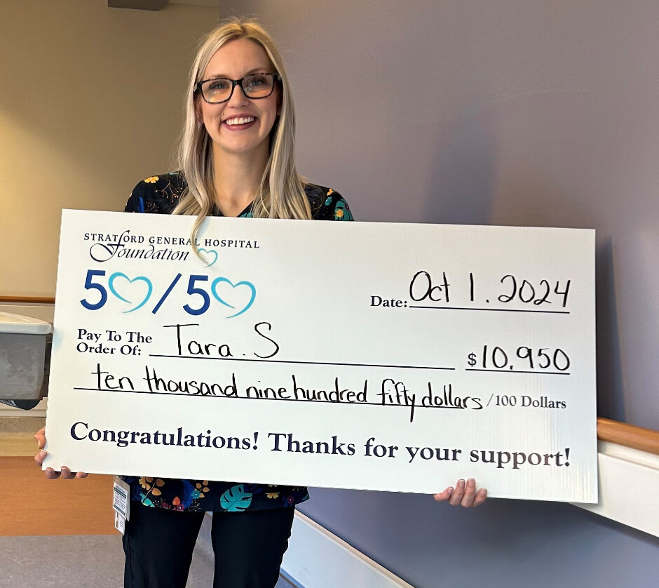 A smiling woman with long blonde hair and glasses holds an oversized cheque indoors. The cheque, from the Stratford General Hospital Foundation’s 50/50 fundraiser, is made out to “Tara S.” for $10,950, dated October 1, 2024, with the message, “Congratulations! Thanks for your support!”